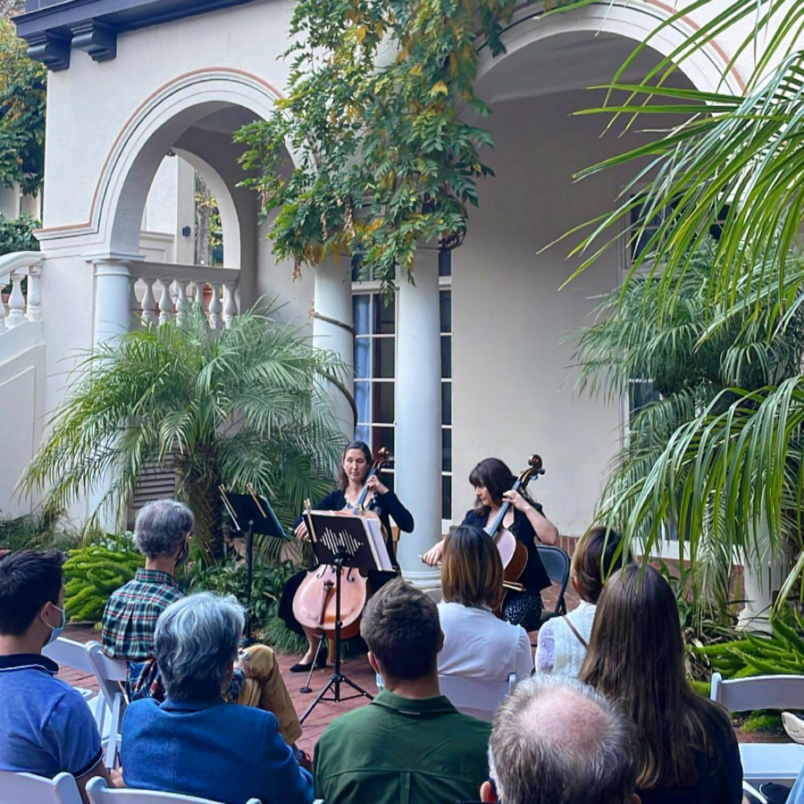 Image: Two cello players perform in a courtyard surrounded by a seated audience